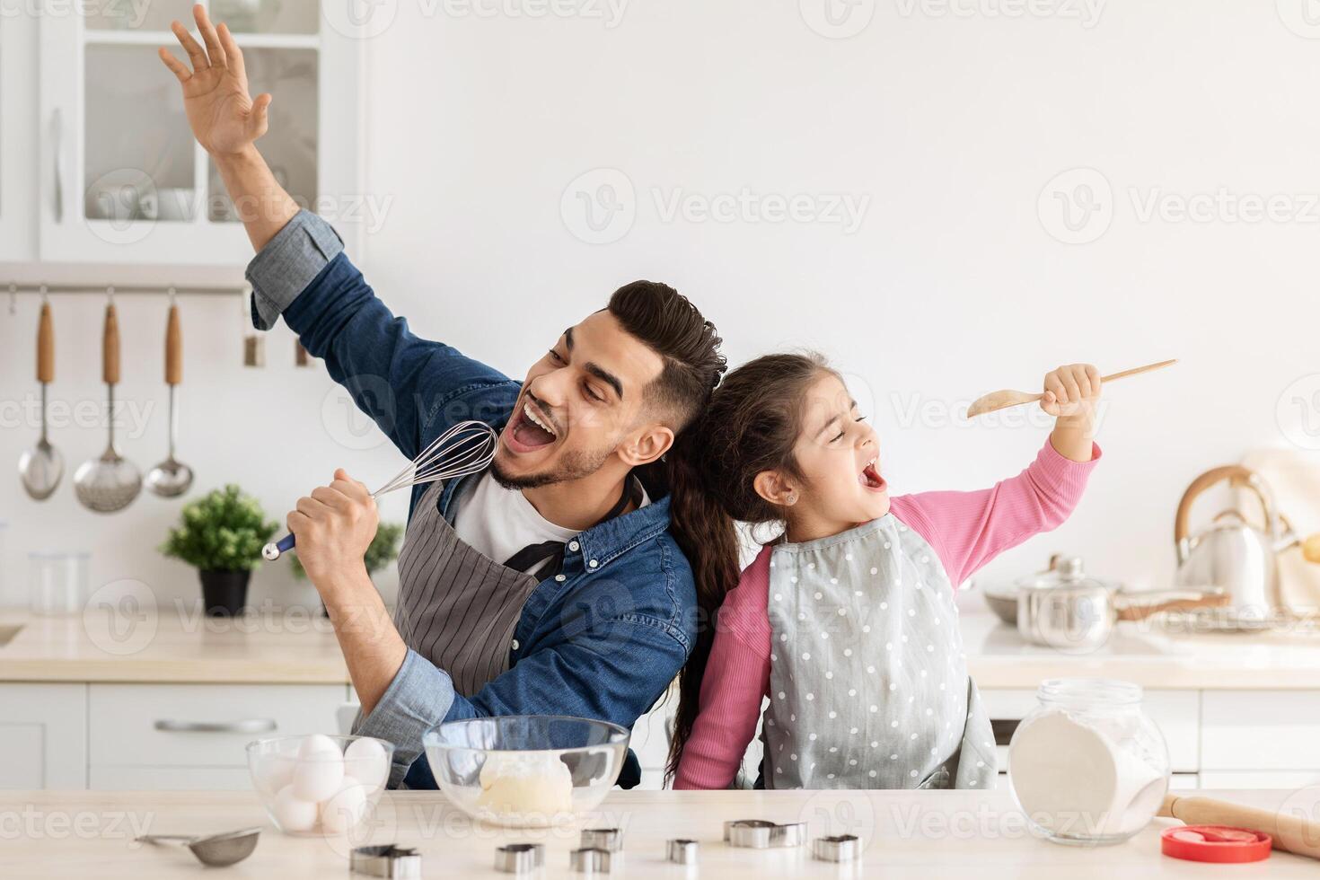 Cheerful arab father and little daughter having fun while baking together in kitchen, happy middle eastern dad and child singing and fooling, using spatula and whisk as microphones, copy space photo
