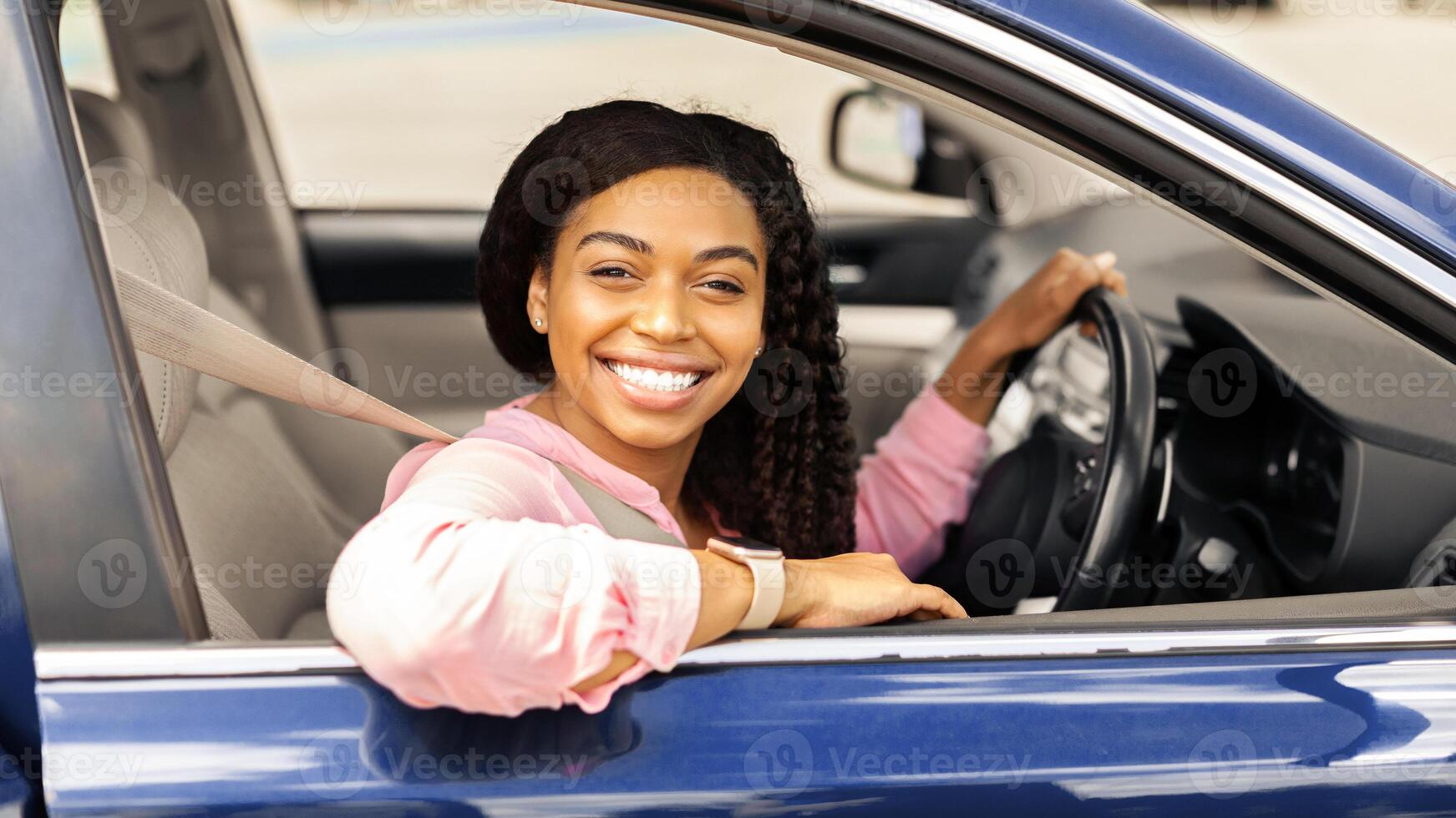 Positive young African American lady sitting on driver's seat. Happy black female posing looking at camera through open automobile door and smiling. Auto test-drive, leasing, buying, rental concept photo