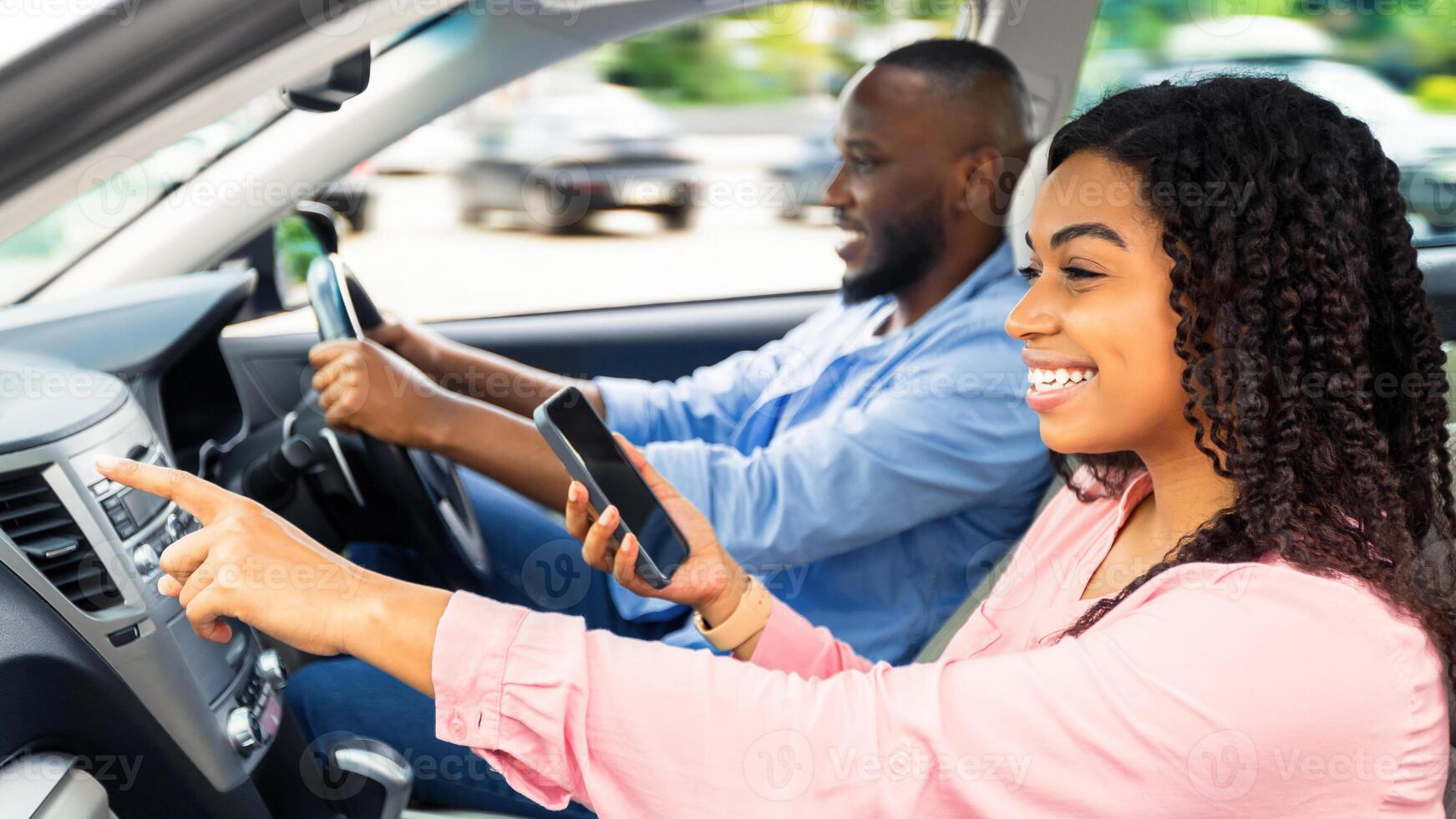 Road Trip Concept. Profile side view portrait of smiling black couple sitting in luxury car, man driving auto, woman with mobile phone pointing at the road, showing way, using digital map application photo