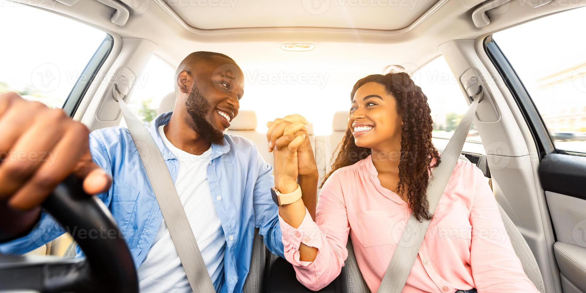 Loving African American couple holding hands and smiling to each other while sitting in comfortable auto, front view. Happy guy having car trip with his beautiful girlfriend, summer vacation concept photo