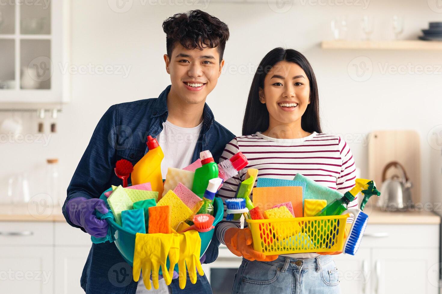 Portrait of smiling asian house-keepers with cleaning supplies posing at camera while cleaning together at home. Cheerful young man and woman enjoying kitchen cleaning, showing tools photo