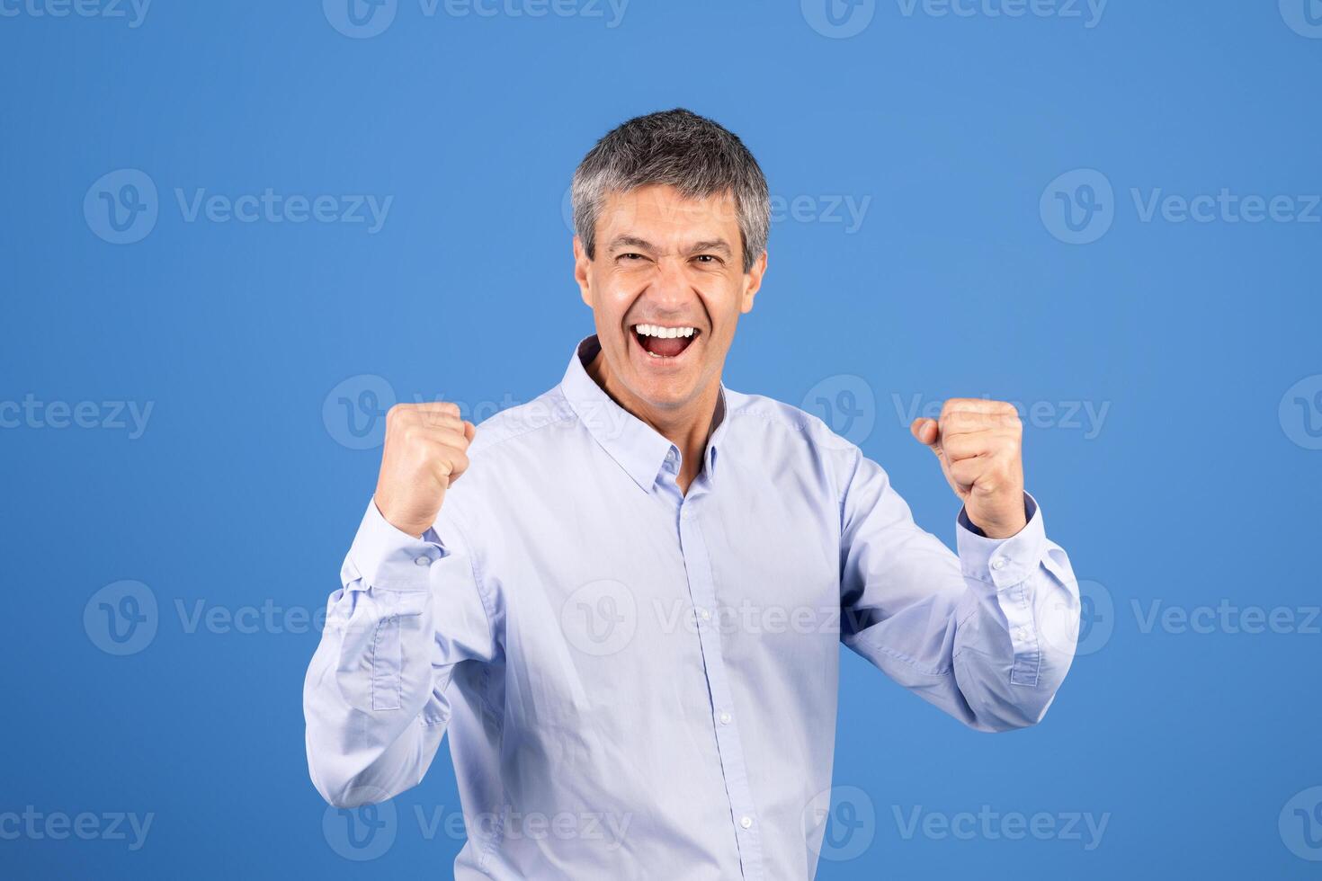 A middle aged European man in his 50s displays excitement and joy with raised fists. He is wearing a light blue shirt and smiling broadly, exuding positivity in a vibrant blue setting. photo