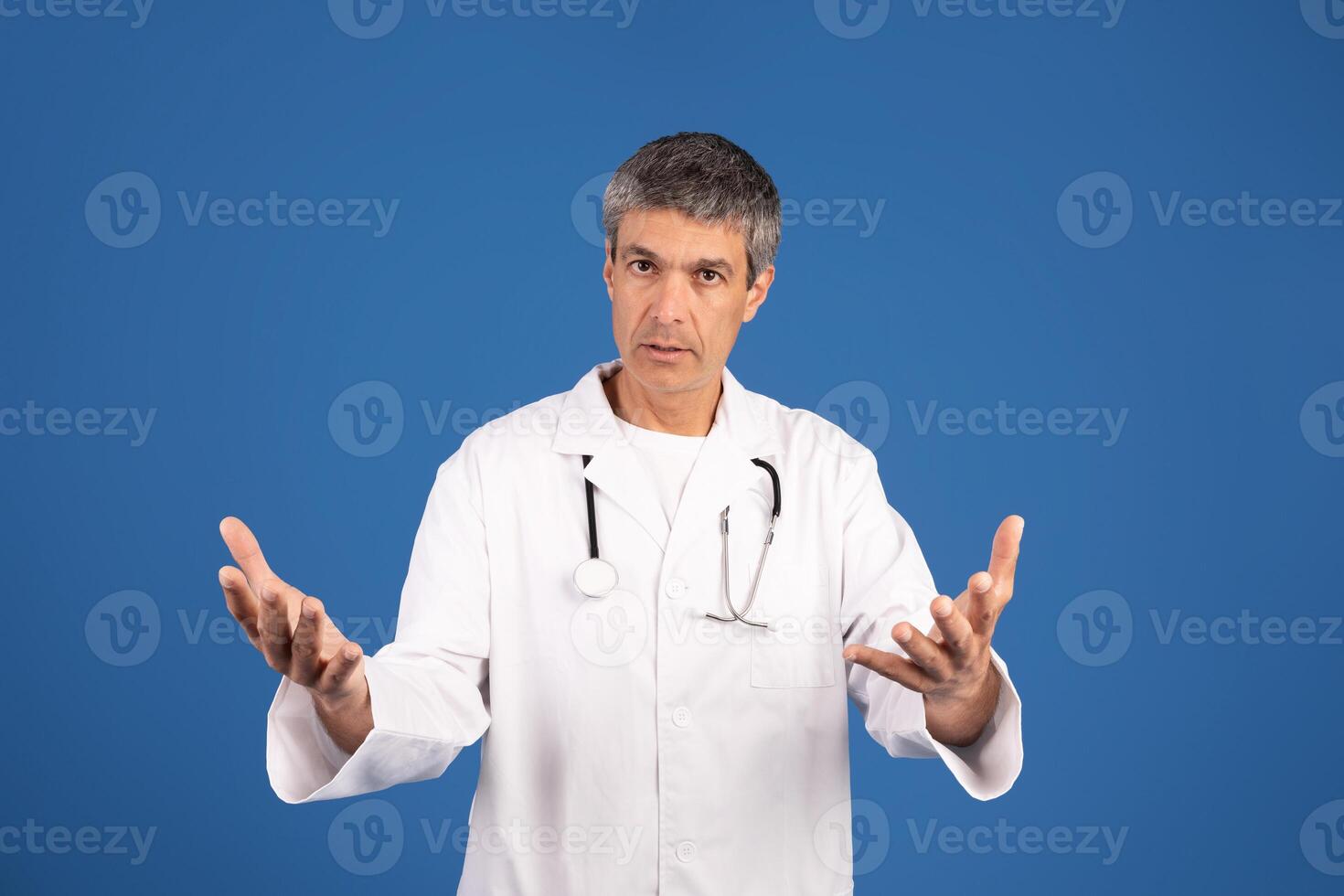 A mature Caucasian man dressed in a white doctor's coat expresses engagement and concern, using open hand gestures to communicate. The plain blue background emphasizes his presence. photo