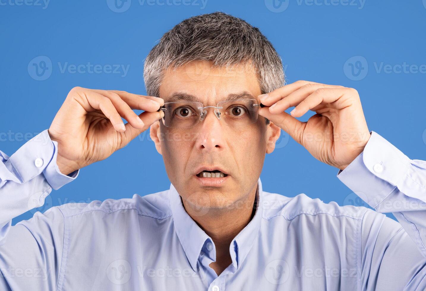 A mature European man in his 50s adjusts his glasses, looking surprised and isolated against a bright blue background. He wears a light blue shirt and appears focused on something in front of him. photo