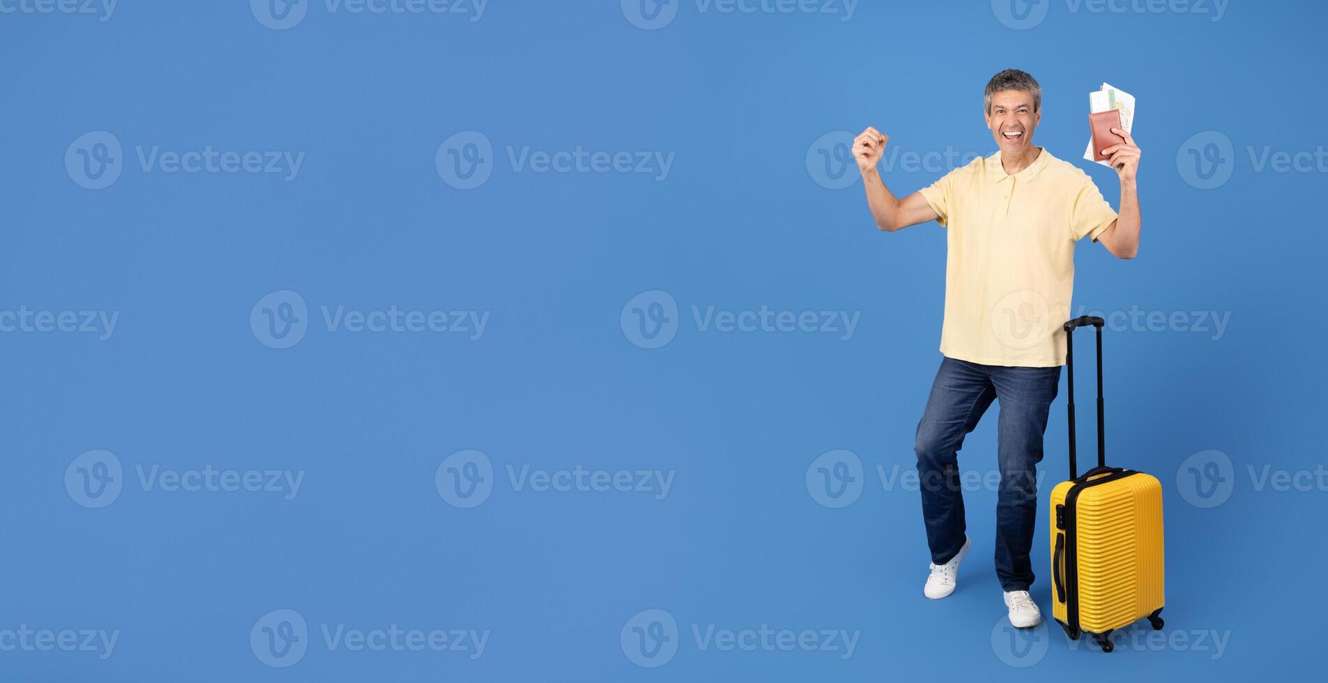 A cheerful middle aged man stands against a blue background, holding up his passport and displaying excitement for his upcoming travels. He has a suitcase beside him, ready for adventure, copy space photo