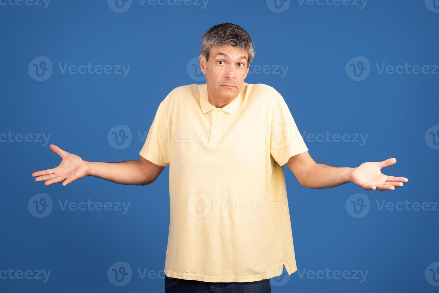 A mature Caucasian man in his 50s stands with a puzzled expression, his arms raised in a shrug. He wears a yellow polo shirt, showcasing a moment of contemplation or confusion. photo