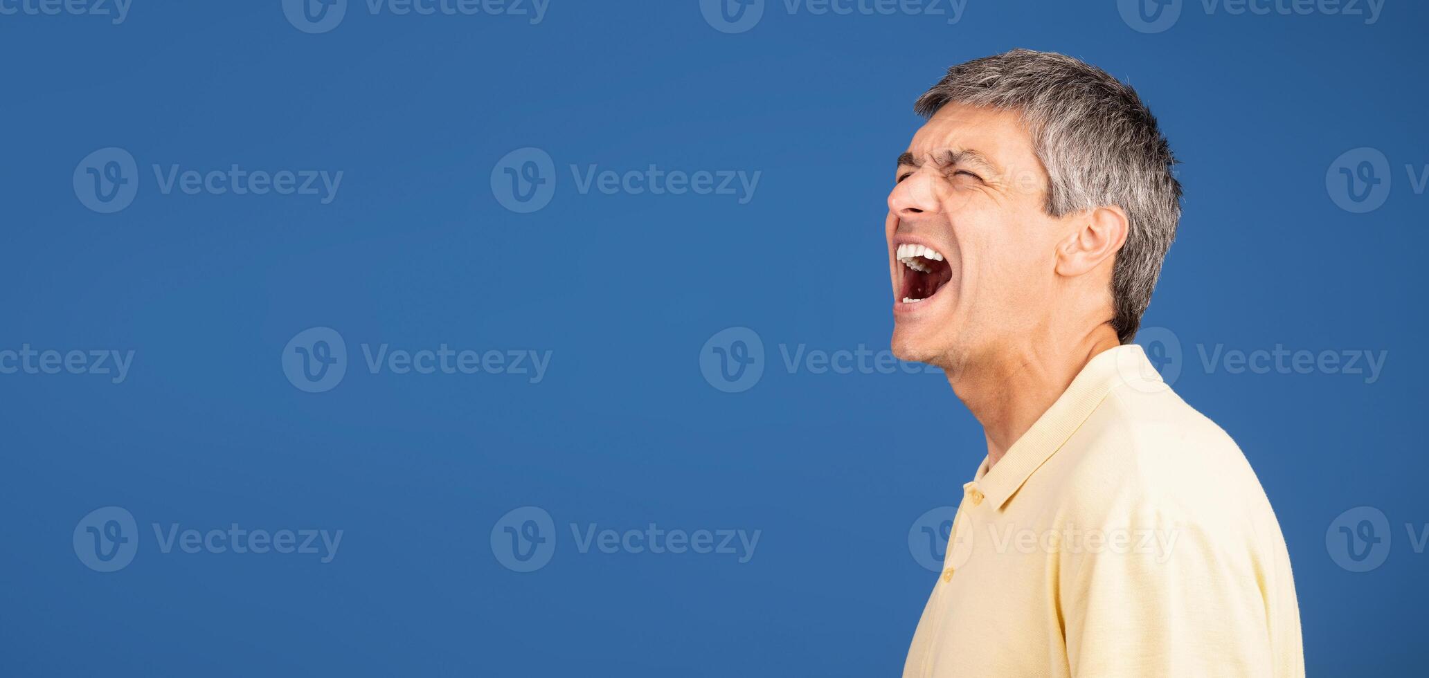 A middle aged man in his 50s yells out in frustration while standing alone against a vibrant blue background, conveying a sense of isolation and emotional intensity, copy space photo