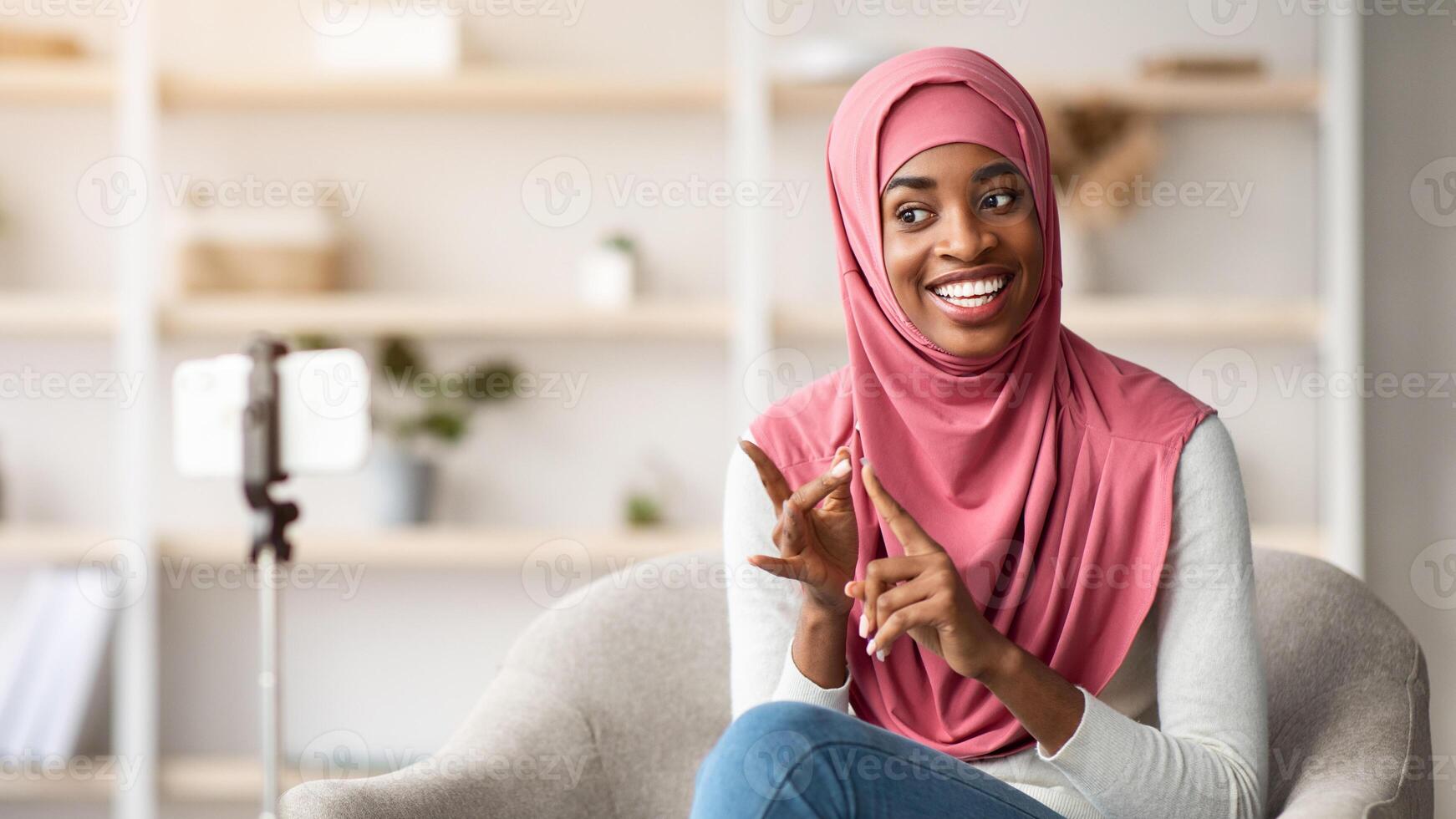 Young black islamic woman in hijab recording new vlog on smartphone at home, smiling african muslim lady making live stream with mobile phone on tripod, talking and gesturing at camera photo