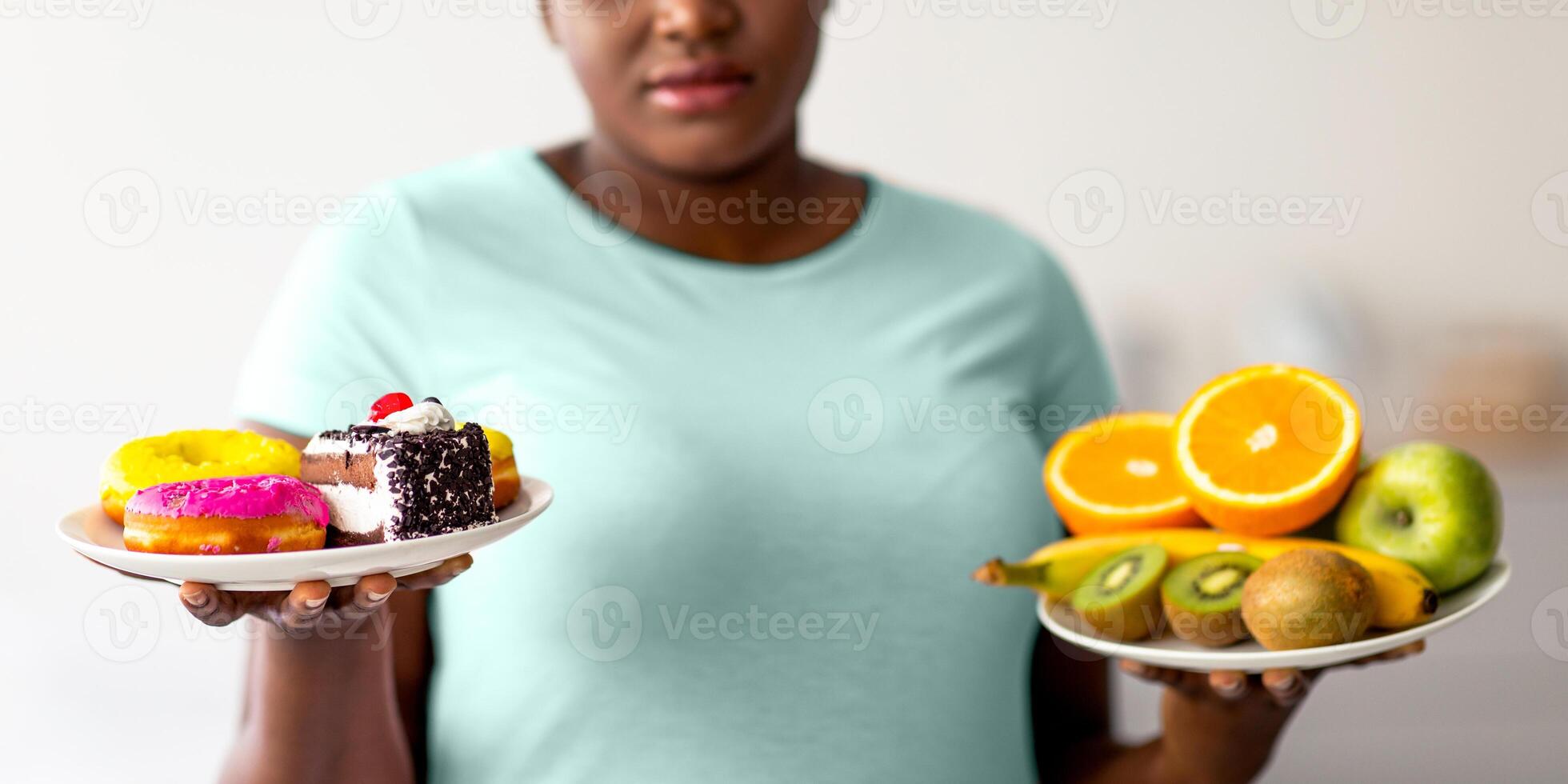 Obese Afro woman holding plates with fruits and desserts, tempted to break weight loss diet at home, selective focus. Overweight young black lady choosing between healthy and unhealthy nutrition photo