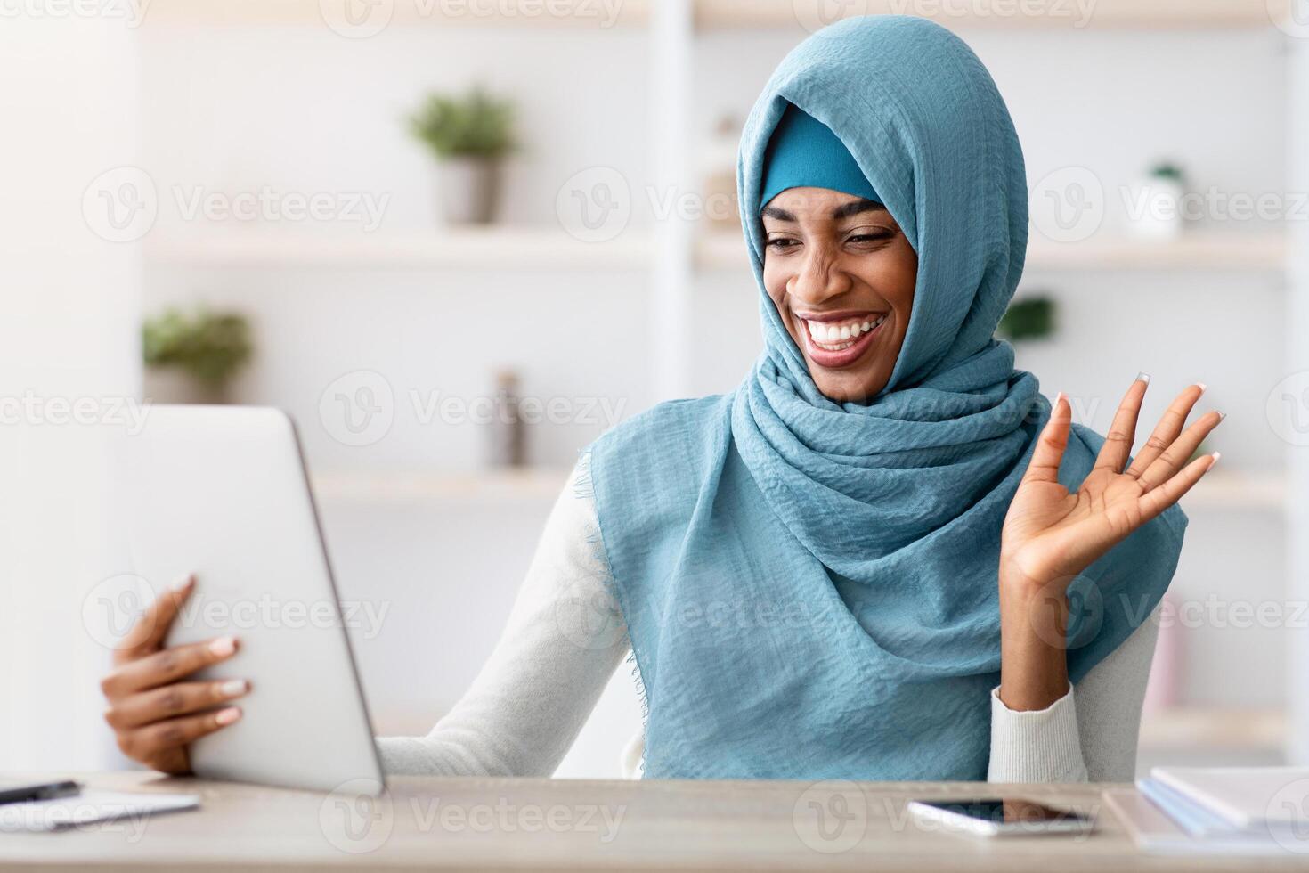 Online Communication. Cheerful Black Muslim lady In Hijab Making Call With Digital Tablet While Sitting At Desk In Home Office, African Islamic Freelancer Lady Waving Hand At Web Camera photo