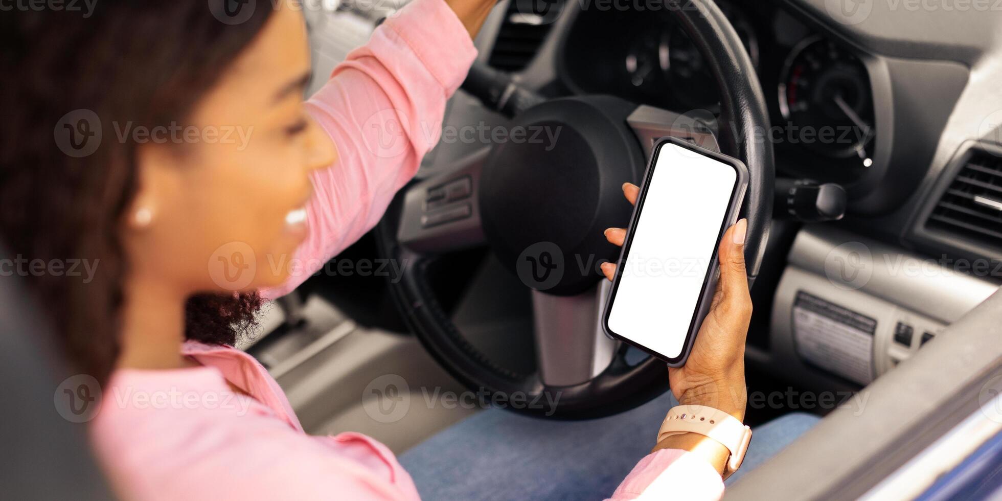 Over the shoulder view of happy black female driver holding smartphone with white blank screen, using mobile app for navigation, mock up. Closeup of cell phone with empty display, copy space template photo