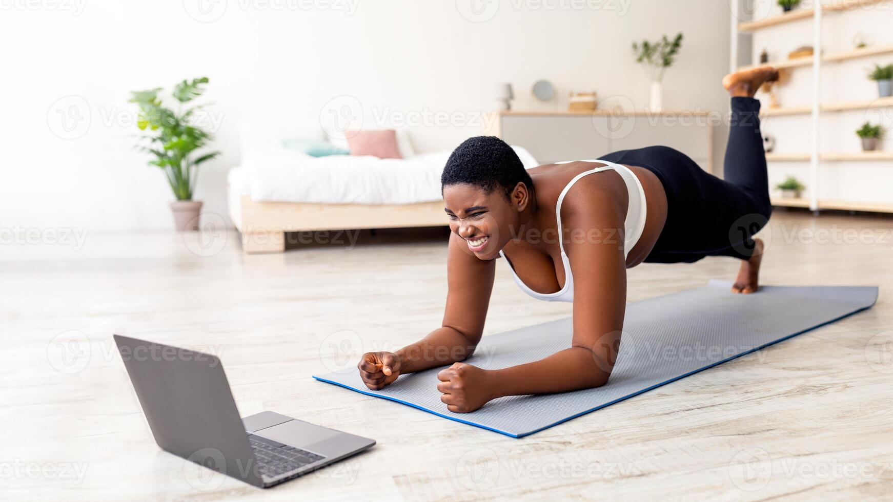 Plus size Afro lady working out at home with online personal trainer, using laptop, standing in elbow plank pose. Overeight black woman training to tutorial on web, indoors photo