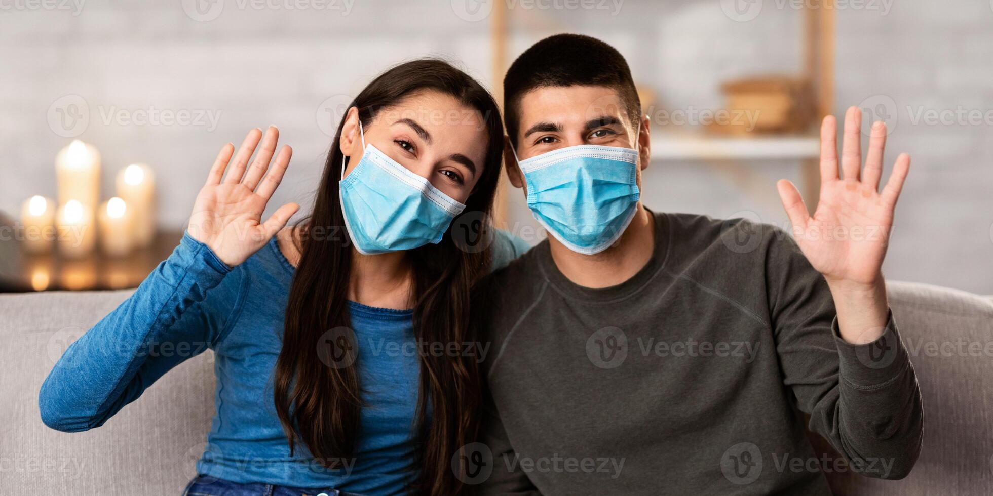 Smiling couple wearing surgical face mask sitting on sofa at home and waving hands to camera, showing hello sign photo