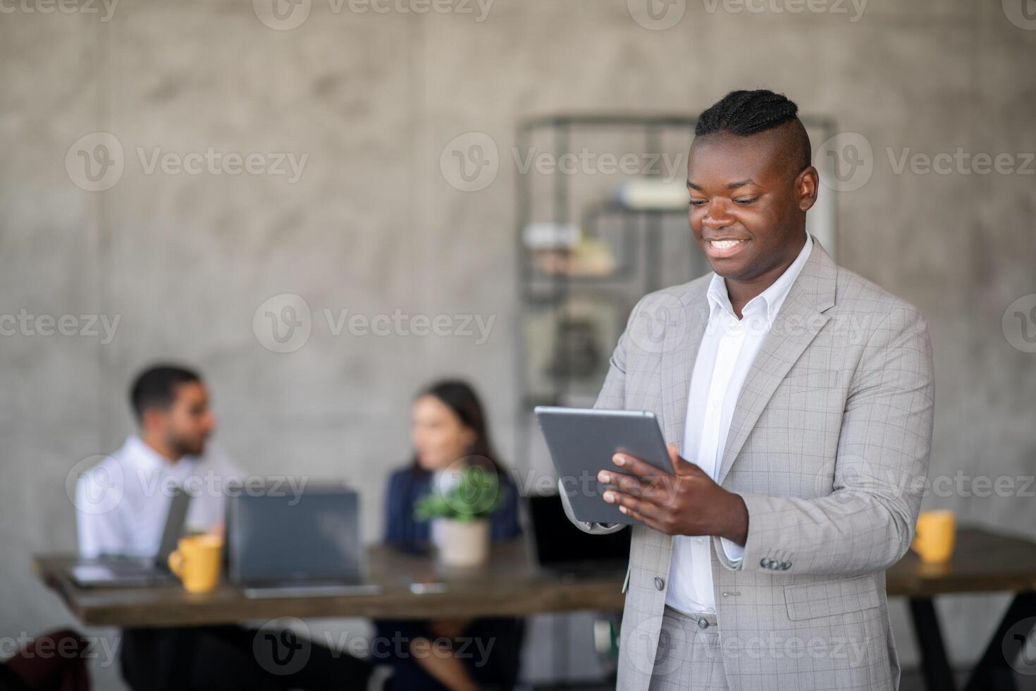 A business professional stands in a stylish office, reviewing information on a tablet while colleagues engage in discussion nearby. The atmosphere highlights teamwork and international cooperation. photo