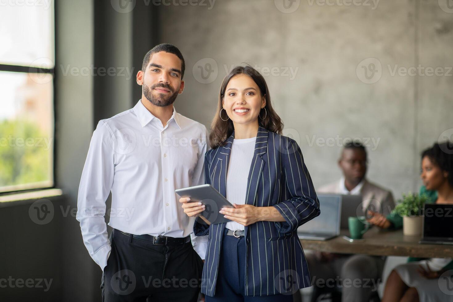 Two professionals stand together in a stylish office, engaging with digital devices. Behind them, a diverse team discusses projects, reflecting teamwork and international cooperation. photo