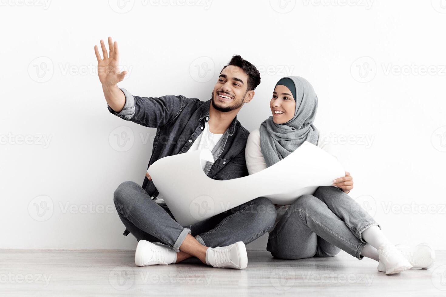 Excited young arab couple working on new home interior and design, happy handsome husband and wife in hijab sitting on floor by empty white wall, holding apartment scheme, looking at copy space photo