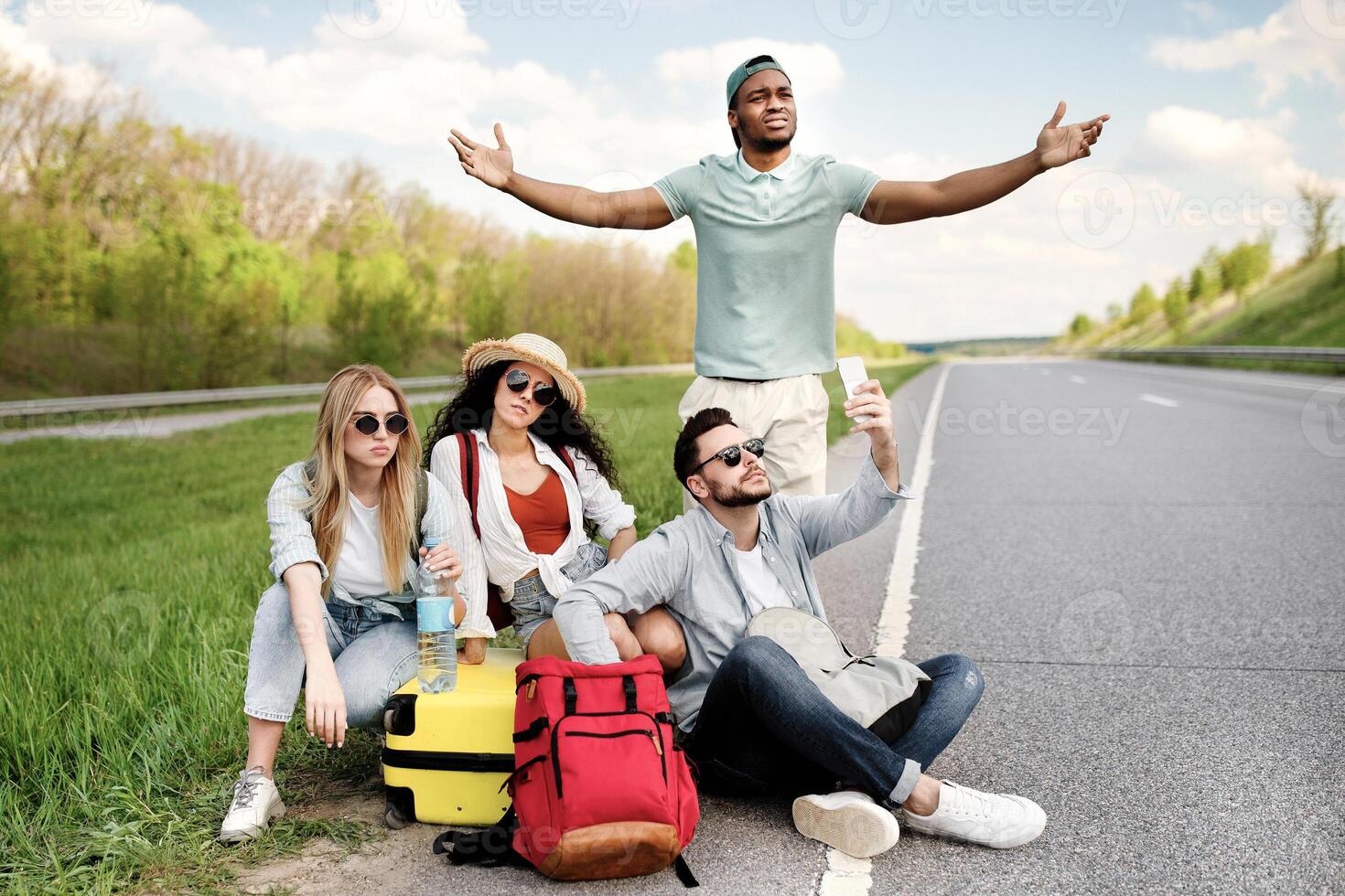 Unhappy millennial tourists sitting on roadside, cannot catch car, waiting for too long, having no smartphone connection. Young friends hitchhiking on highway, having problem finding free ride photo