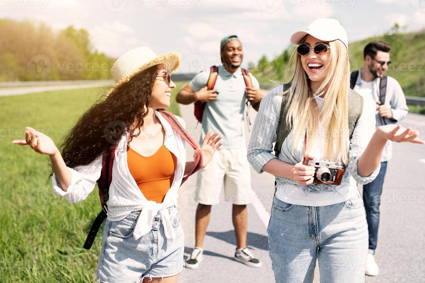 Group of multinational millennial friends walking along highway, chatting to each other, laughing, having fun. Hitchhiking, summer adventure, autostop travel, tourism concept photo