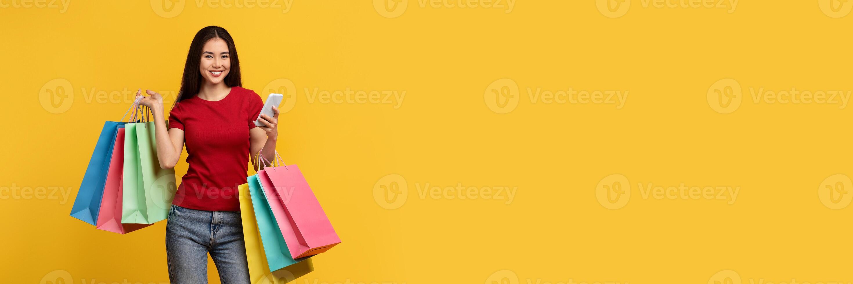 Online coupons, discounts, shopping mobile app. Happy attractive young asian woman wearing casual clothing carrying shopping bags, checking smartphone, copy space, yellow background photo