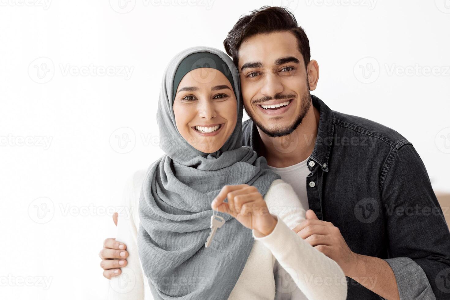 Happy young arab family husband and wife embracing and showing apartment key, copy space. Handsome middle-eastern man hugging his beautiful woman in hijab, moving to new house, relocation concept photo