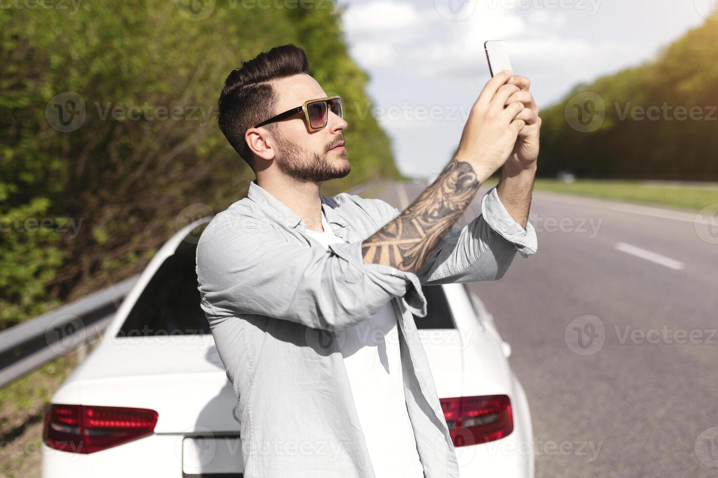 Upset millennial man standing on road next to broken auto, raising hand with cellphone, trying to call roadside assistance. No connection, car breakdown, bad mobile network concept photo