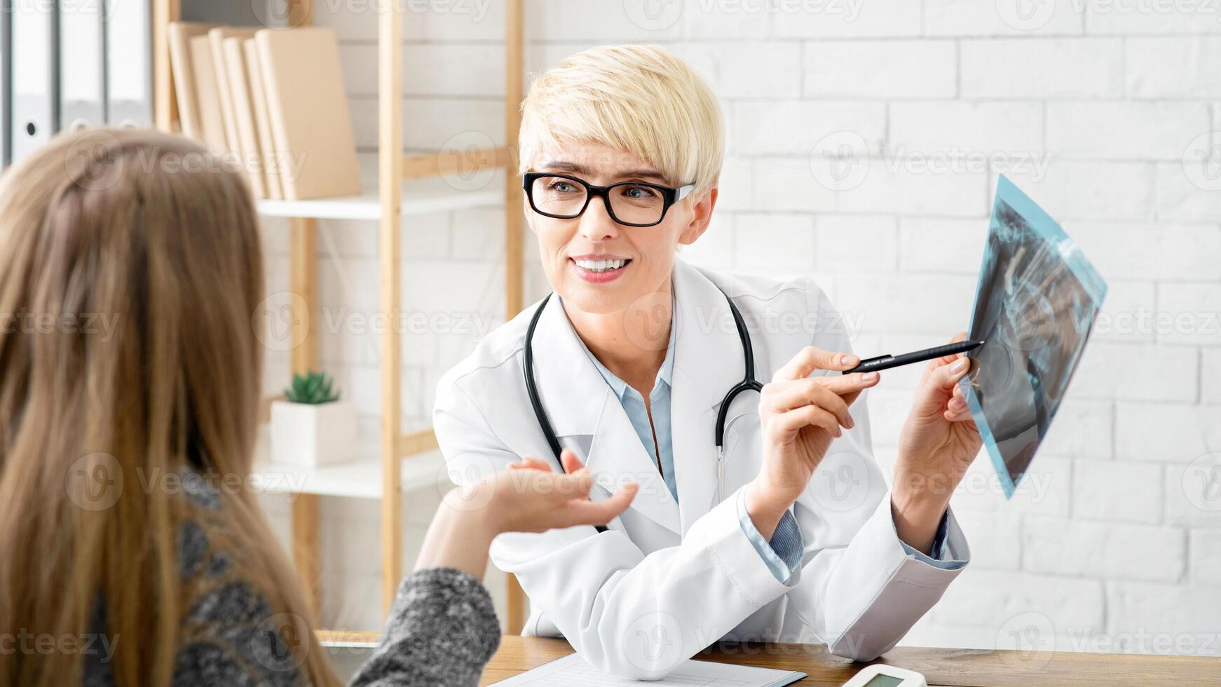 Consultation with doctor in clinic. Adult female therapist in coat and glasses shows x-ray and explains problem to patient in modern office interior at table with tablet and tonometer, free space photo