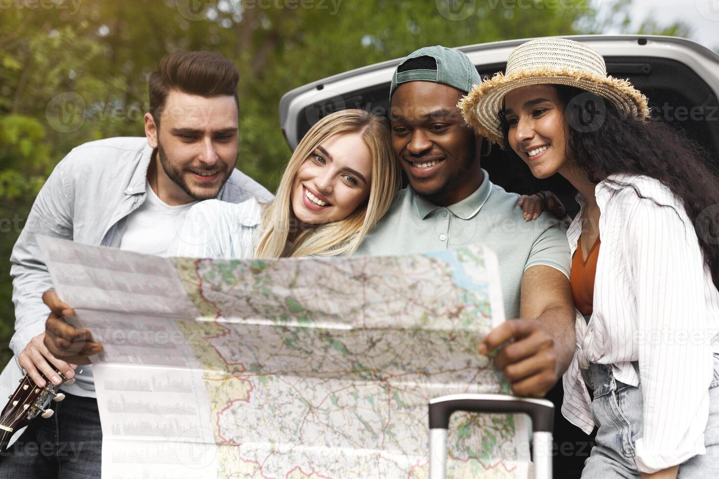Group of multiracial young friends looking at map while sitting in open car trunk on road, traveling together in countryside. Millennial students having fun trip, hitchhiking on summer vacation photo