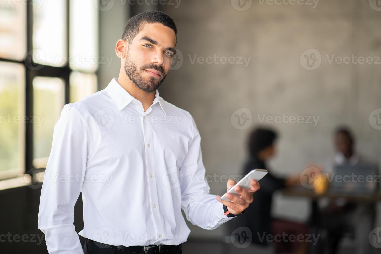 A confident man in a white shirt holds a smartphone while standing in a corporate office. In the background, a diverse team collaborates on a project, reflecting active teamwork. photo