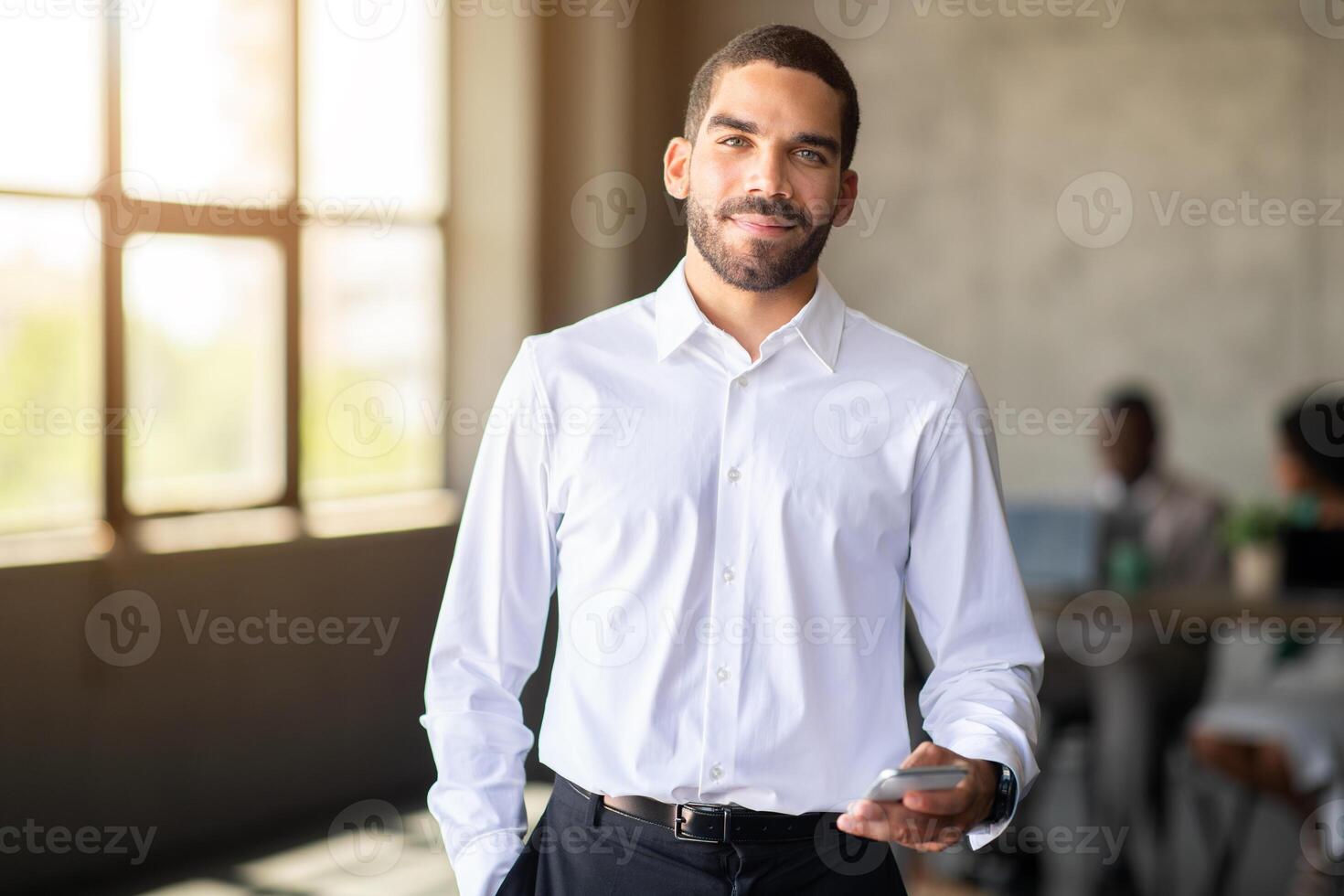 A confident man in a white shirt holds a smartphone while interacting with his diverse team in a corporate office. The atmosphere is focused on teamwork and achieving success during their meeting. photo