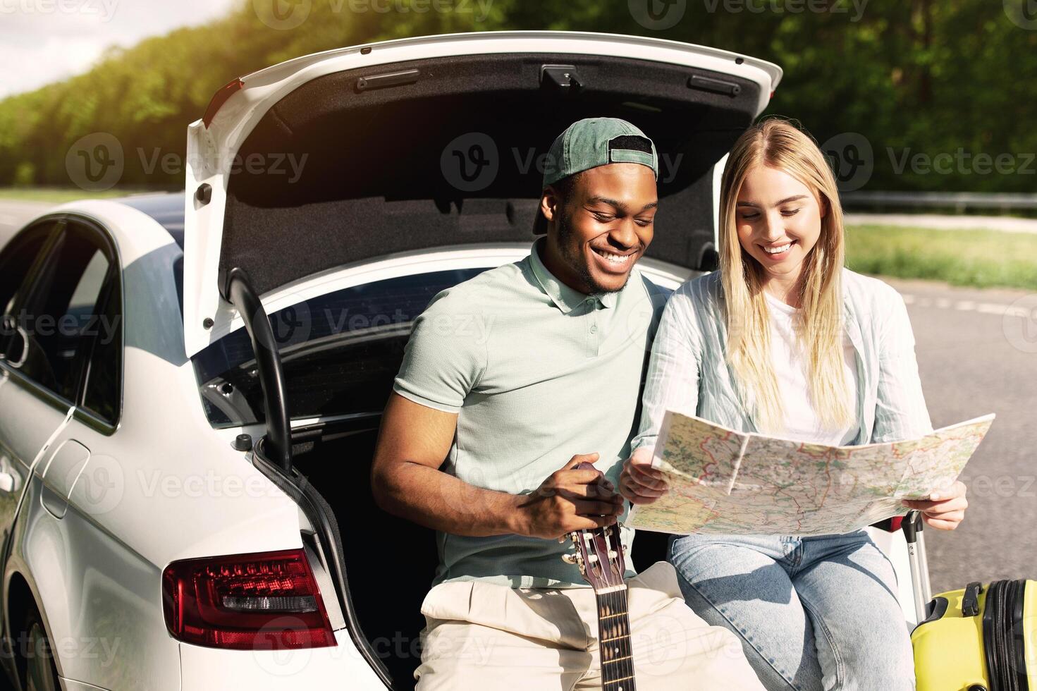 Millennial multiracial couple going on road trip in car, looking at map, taking break from driving, sitting in auto trunk together. Young black guy and his girlfriend having summertime journey photo