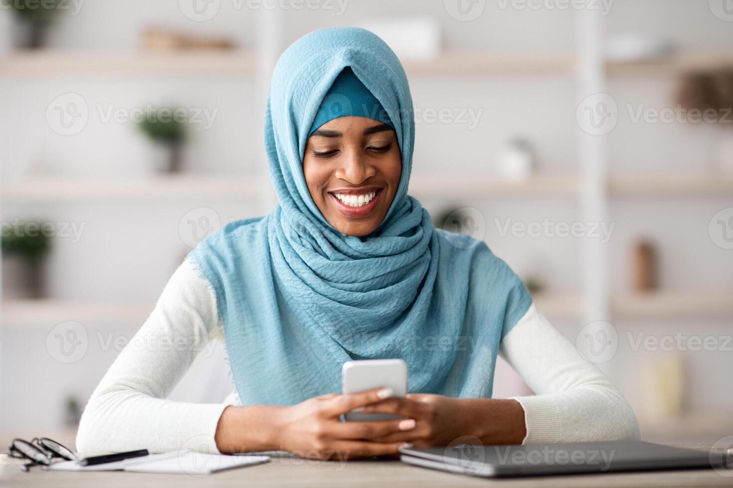 Portrait Of Smiling African Islamic Lady In Hijab Using Smartphone In Office, Cheerful Black Muslim Woman Sitting At Desk With Cellphone In Hands, Browsing Internet, Shopping Online Or Messaging photo