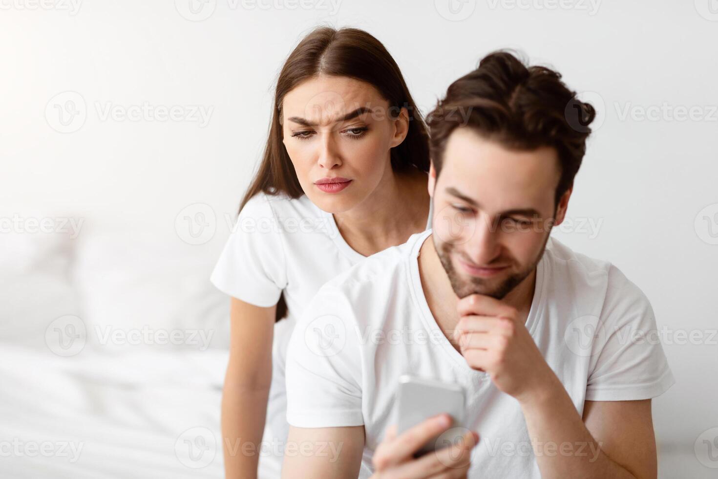 Jealousy. Suspicious Wife Reading Husband's Messages On Smartphone Standing Behind His Back While He Texting In Social Media Indoors. Jealous Girlfriend Checking Boyfriend's Chats Suspecting Affair photo