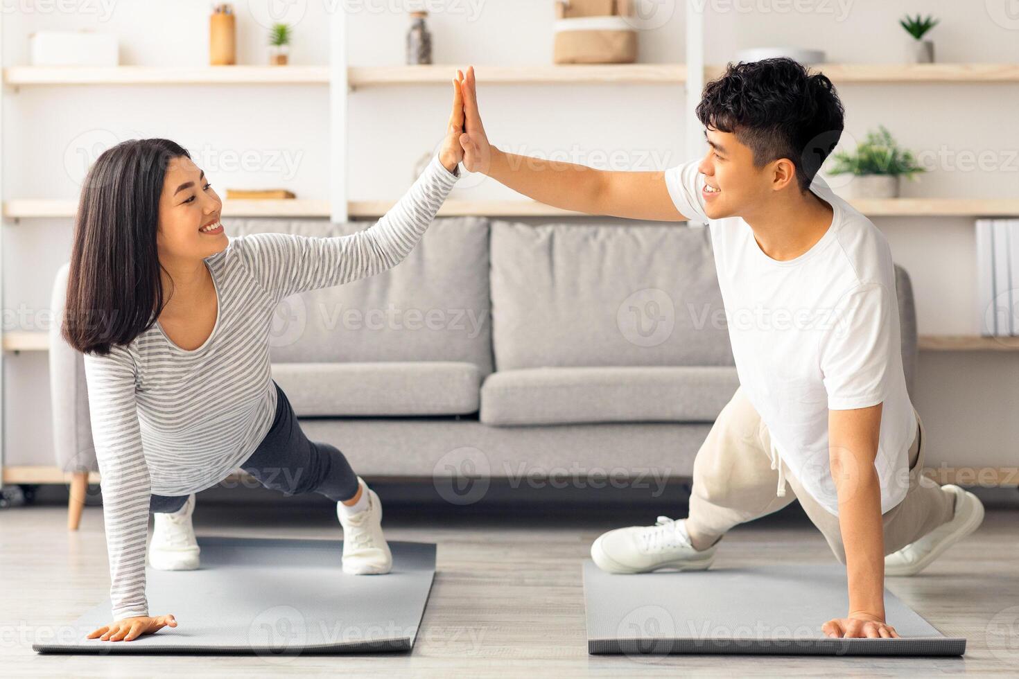 Joyful millennial asian man and woman in sportswear cheering up each other while planking at home, giving high five and smiling, doing exercises on fitness mats at living room, healthy lifestyle photo
