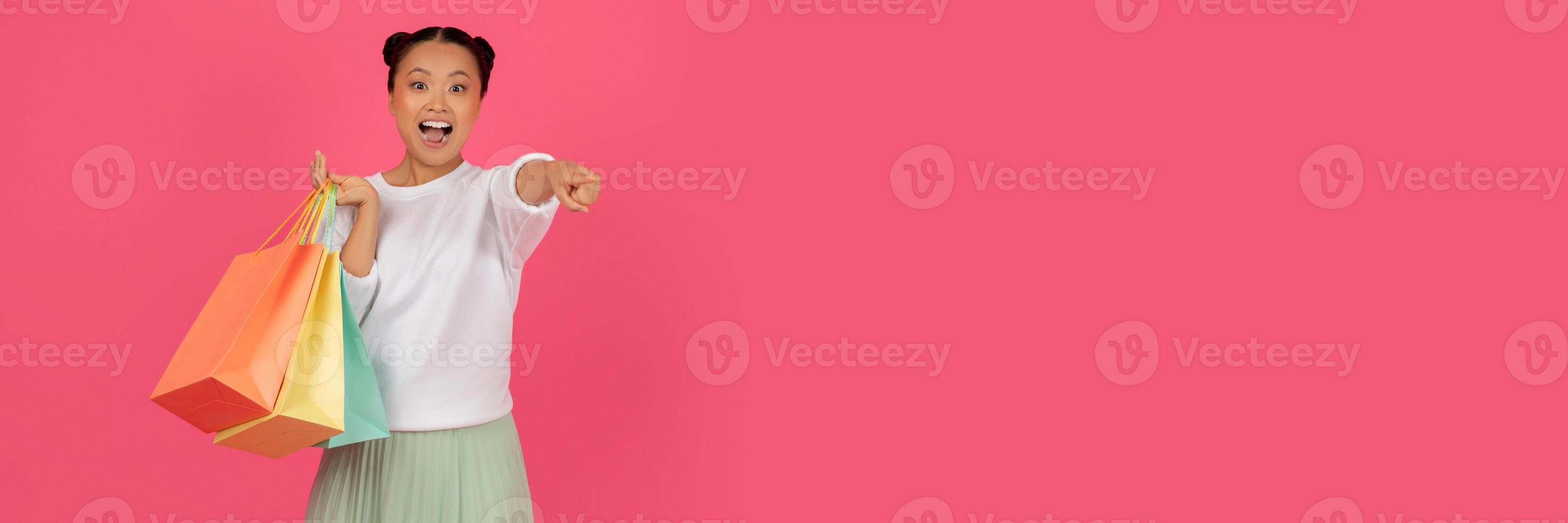 Cheerful young asian lady with shopping bags pointing at camera, cheerful korean woman with purchases selecting you, standing over pink studio background, enjoying seasonal sales, copy space photo