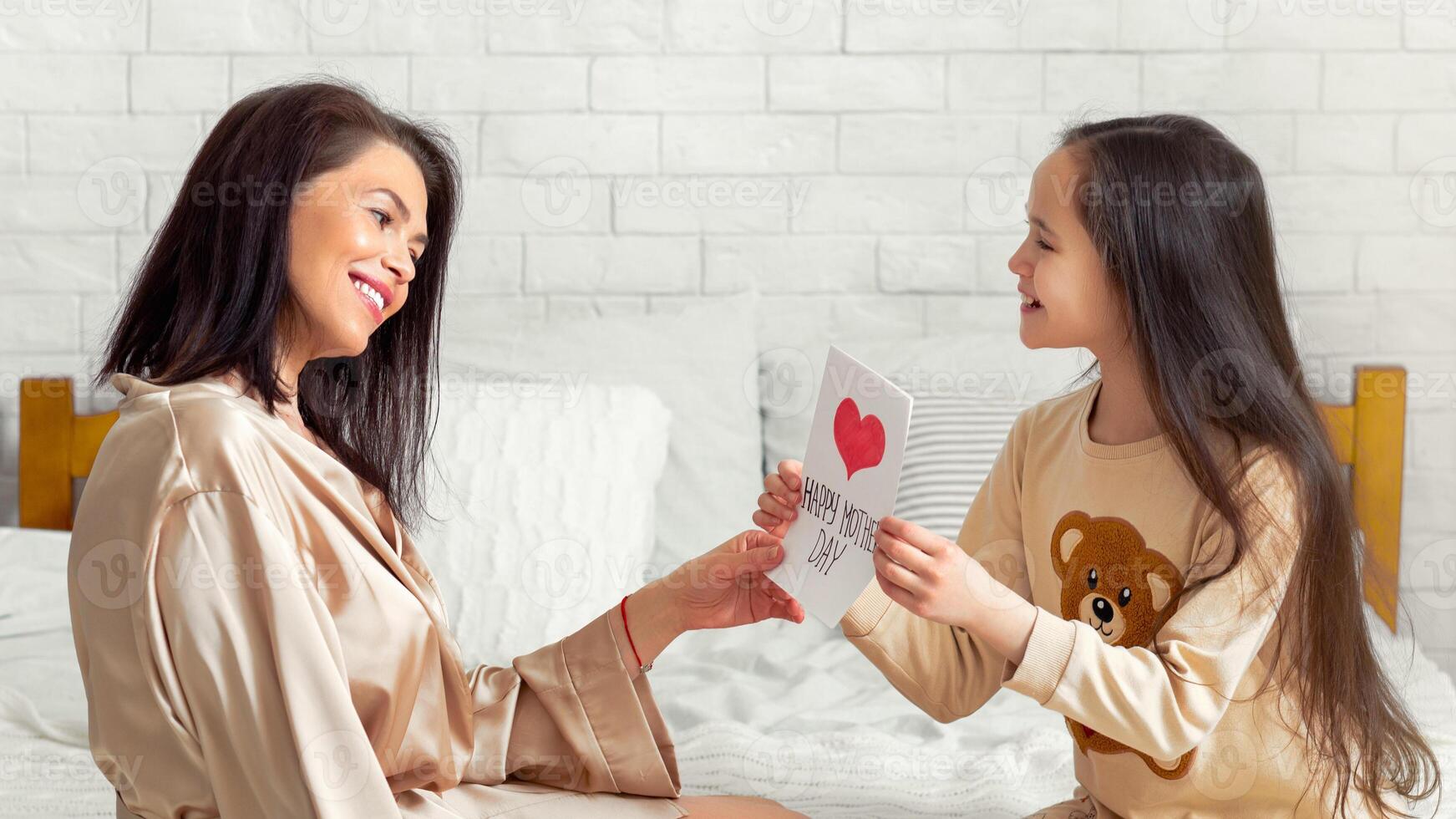 Mother's Day celebration. Cute teen girl giving her mom holiday greeting card on bed at home. Loving daughter congratulating her mummy in bedroom. Family traditions and festivities photo