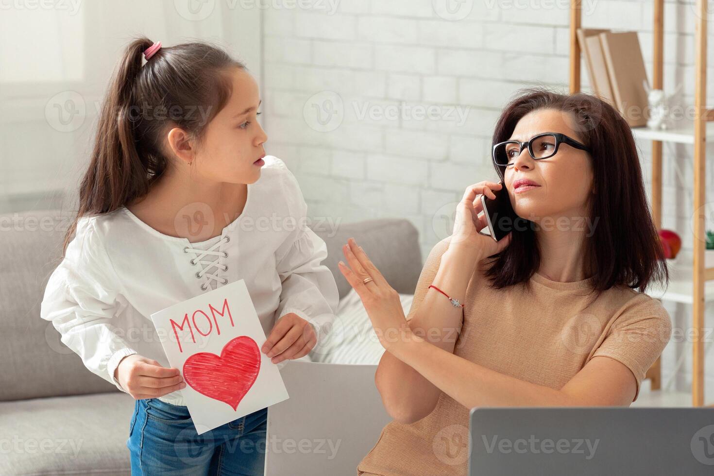 Teen girl giving festive card to her working mother, parent being too busy to pay attention. Business woman having no time for her child, doing online job during coronavirus lockdown photo