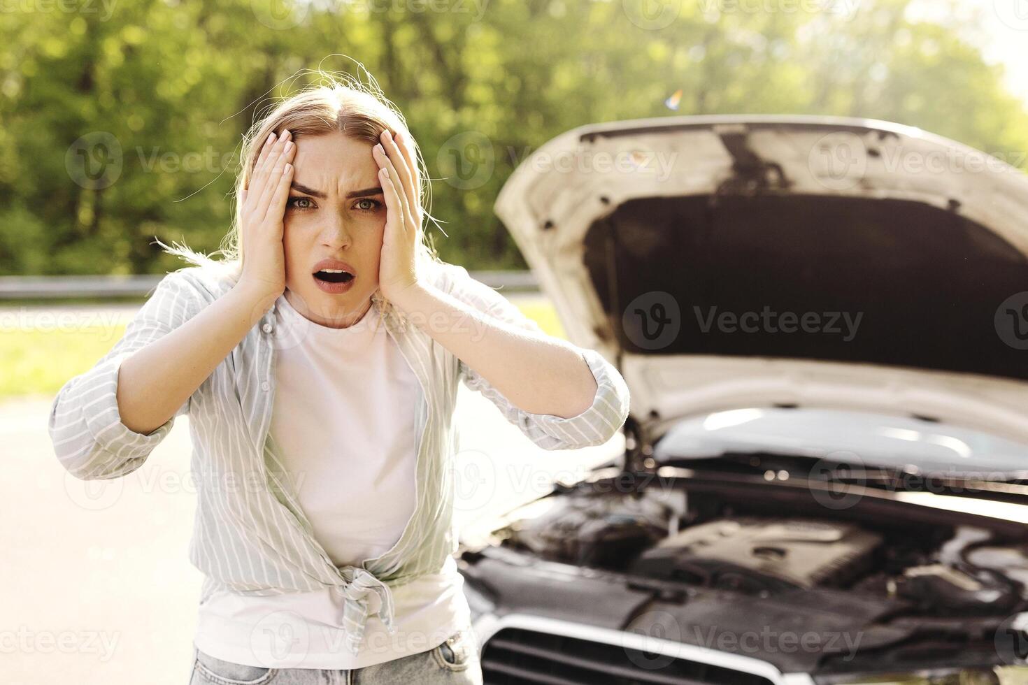 Emotional young blonde woman standing near broken car with open hood, grabbing her head in terror, having road accident. Auto emergency service, automobile breakdown, transport trouble concept photo