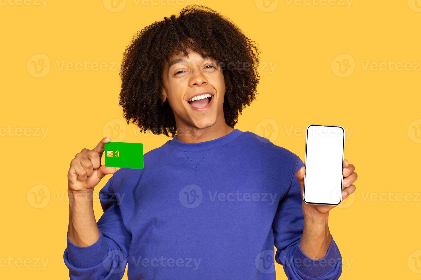 A Brazilian man with curly hair smiles while holding a smartphone in one hand and a green card in the other. The bright yellow background highlights his energetic expression and casual style. photo