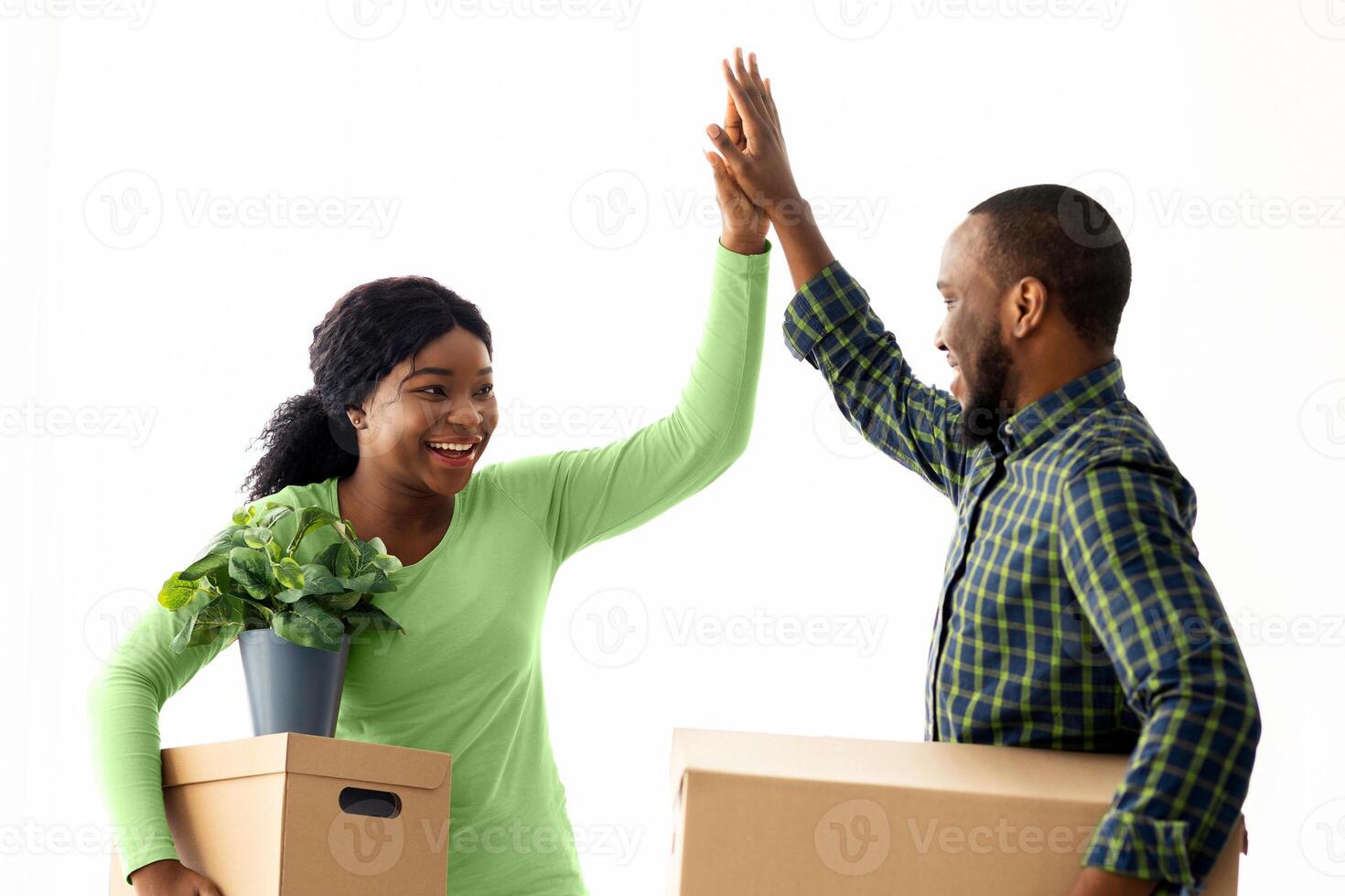 Happy Young Black Couple Giving High Five To Each Other, Celebrating Moving To New House. African American Spouses Carrying Cardboard Boxes And Clasping Hands, Enjoying Relocation, Free Space photo