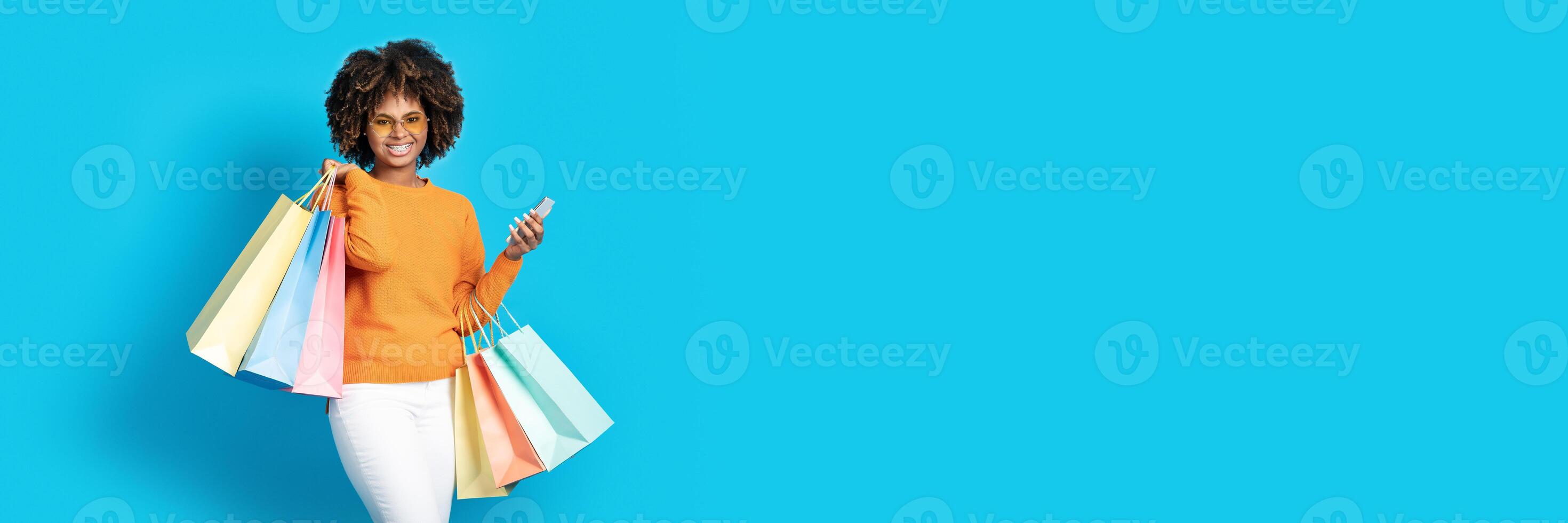 A joyful shopper black woman celebrates a successful Black Friday event, holding colorful bags and checking her phone. Bright turquoise background adds a fun touch to the shopping experience. photo