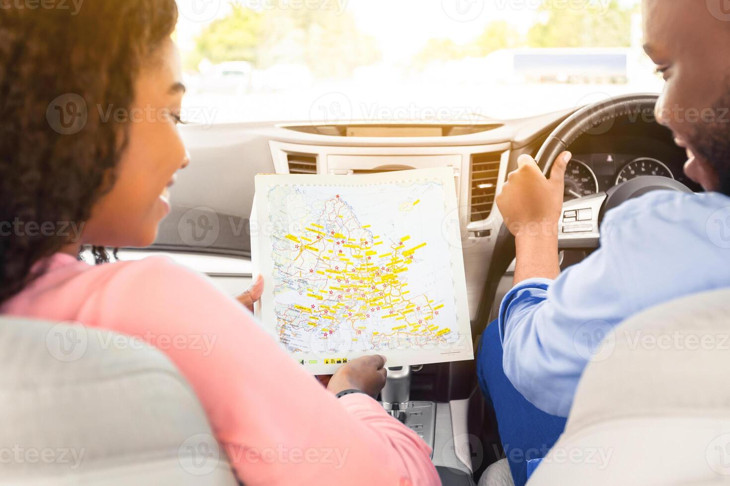 Enjoying Travel Concept. Back view of smiling African American couple holding and looking at paper map sitting inside luxury car, choosing best way to their trip destination, sun flare photo
