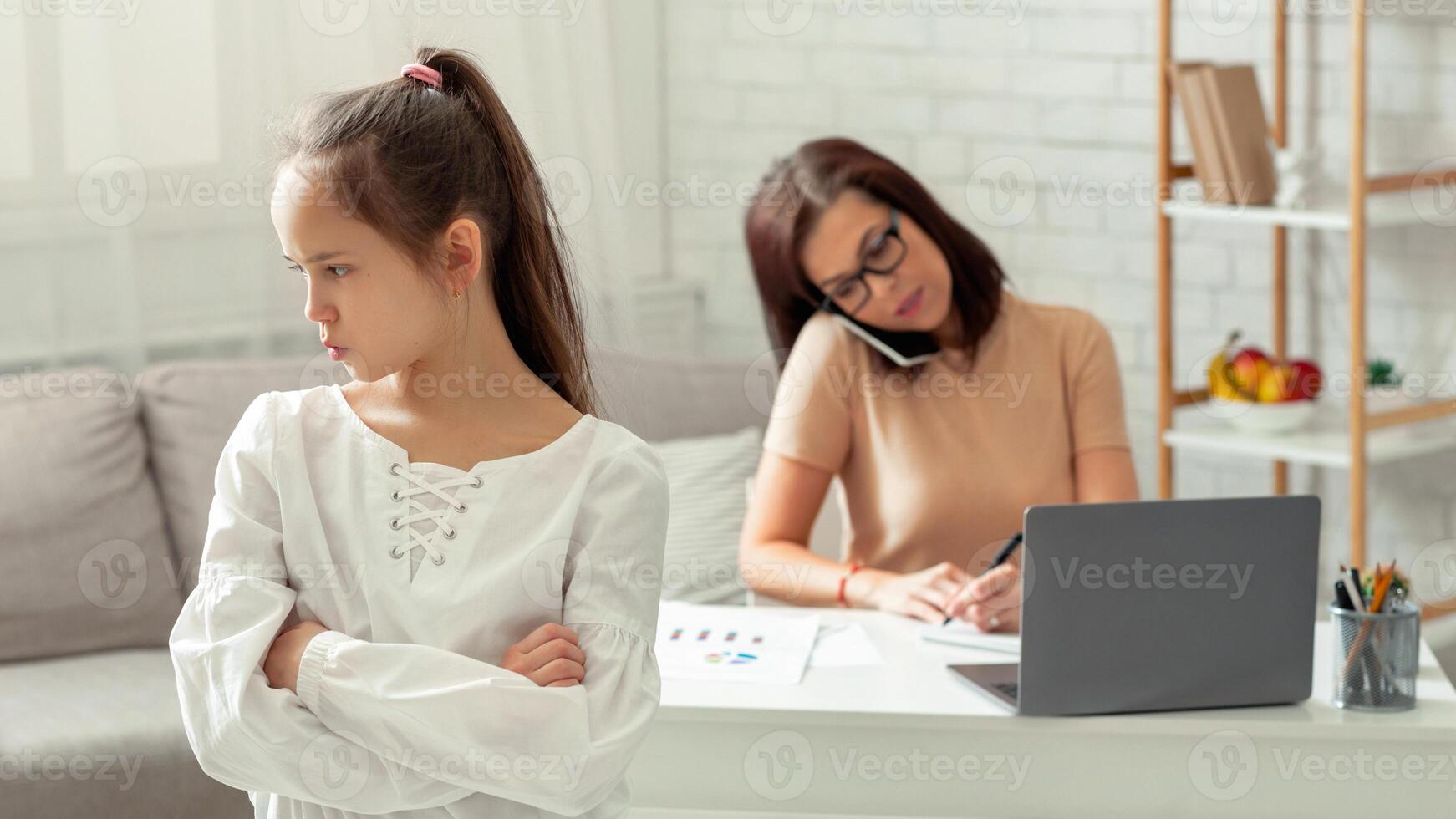 Angry sad teenage girl taking offence at her busy mother working online from home, empty space. Child feeling neglected and lonely with her mom having remote job during covid-19 lockdown photo