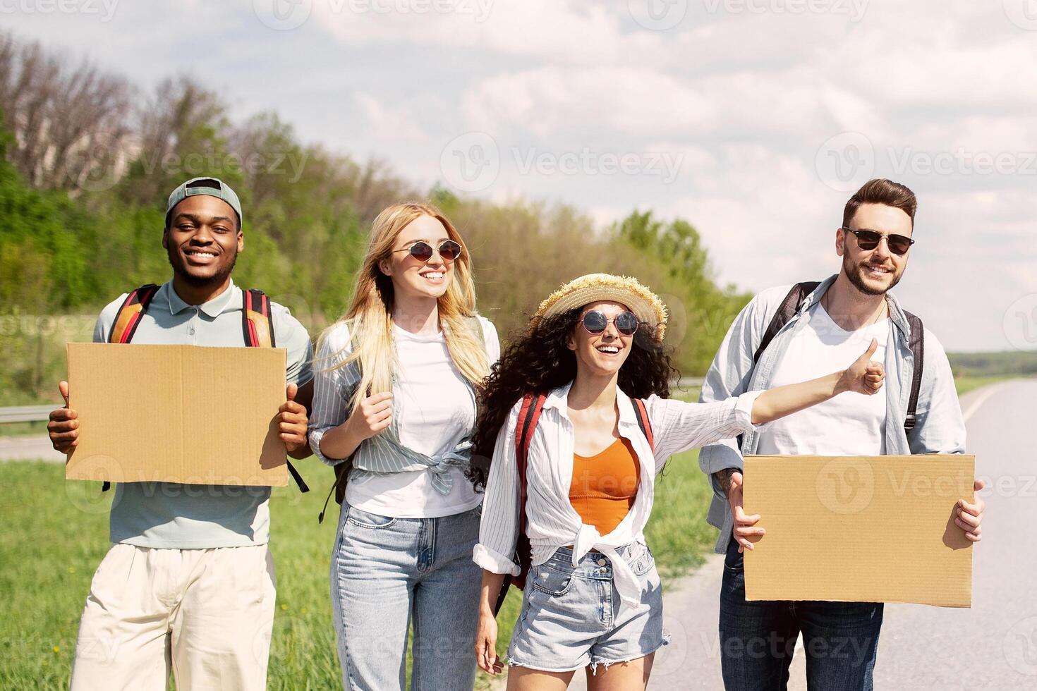 Group of happy multiethnic friends with empty signs hitchhiking on road, catching ride, traveling by autostop, copy space. Cheerful young students going on summer trip together, having fun adventure photo