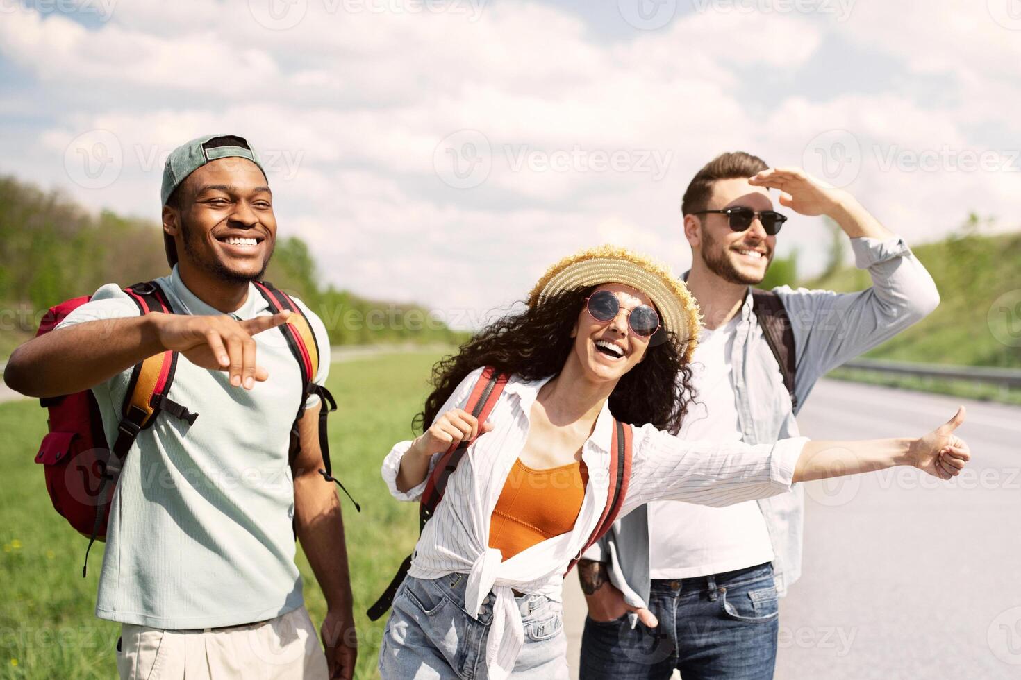 Exciting travel adventure. Diverse friends standing on roadside, stopping cars, hitchhiking for free ride, enjoying summer vacation, copy space. Young multiracial people having autostop journey photo