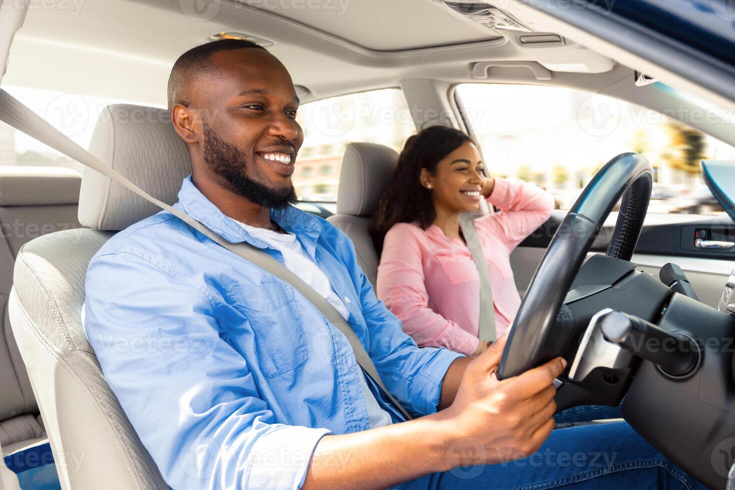 Enjoying Travel Concept. Smiling handsome African American man driving modern luxury car, happy beautiful young woman sitting on the front passenger seat, looking on the road, going on road trip photo