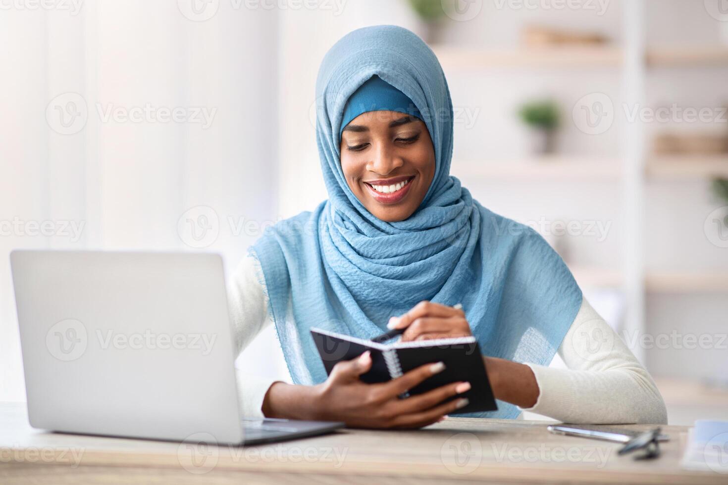 Black Muslim Freelancer Lady In Hijab Taking Notes To Notepad While Working With Laptop Computer At Desk In Home Office, Young African Islamic Woman Planning Business Schedule, Free Space photo