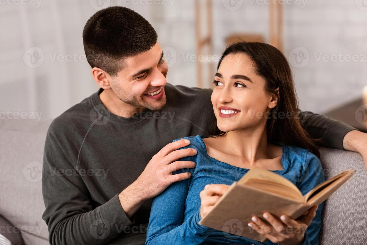 Free Time Concept. Happy spouses sitting on couch, cuddling, reading book together, looking at each other photo