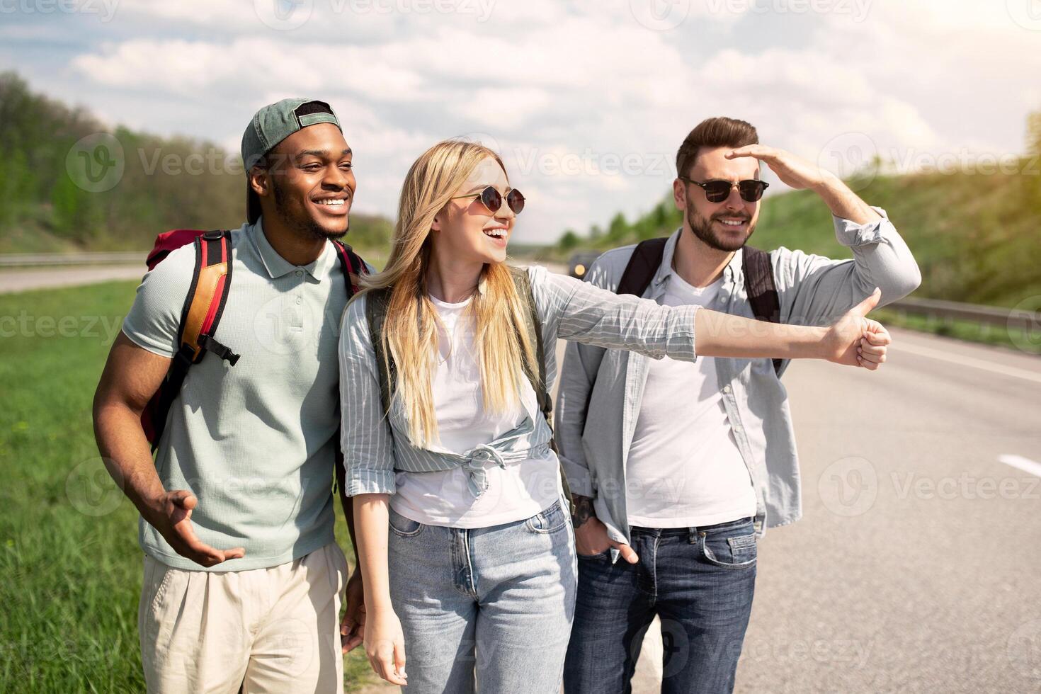Cheerful diverse friends trying to get free ride, hitchhiking on road, showing thumb up outdoors. Free young people traveling by autostop, having cool summer vacation, hitching car on highway photo