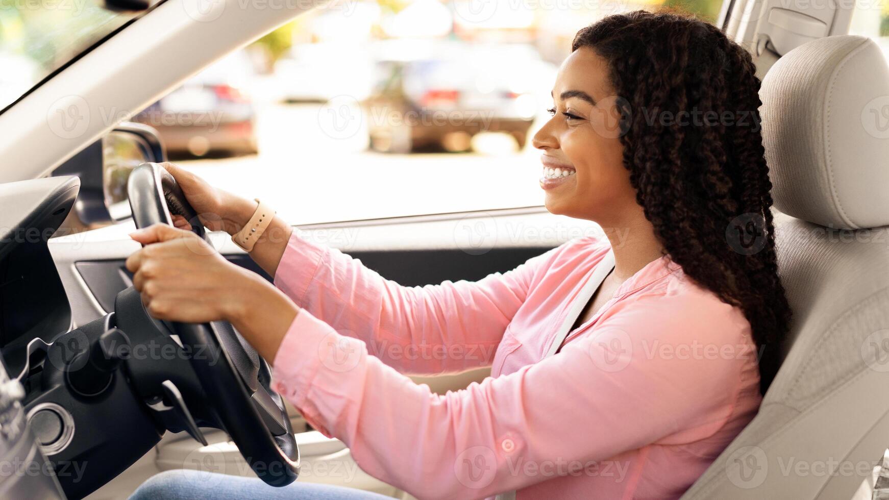 Side view profile portrait of joyful African American woman driving car, doing test drive in dealership showroom. Happy lady riding brand new nice car, sitting on driver's seat in luxury automobile photo