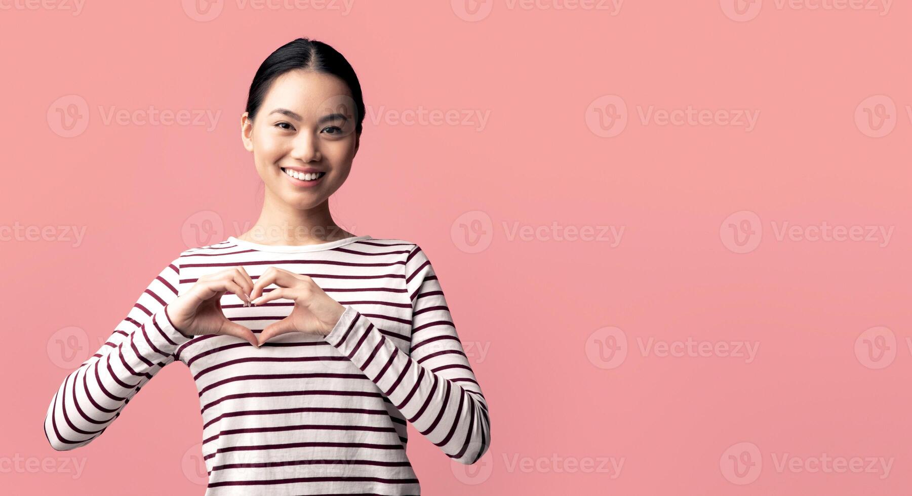 Cheerful young asian lady showing heart gesture with hands near chest, smiling korean woman making love sign and looking at camera, kind millennial female standing on pink background, copy space photo